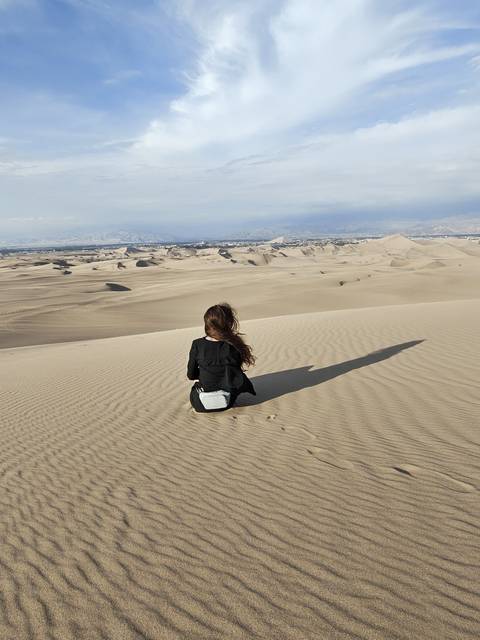       Woman sitting on sweeping golden sand dunes looking over a vast desert landscape with long shadow.
  