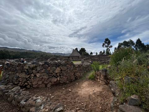       Stone enclosures and a conical thatched structure under a moody sky at an archaeological site.
  