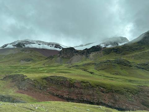       Green pastures and red earth rise to snow-streaked peaks hidden by low clouds.
  