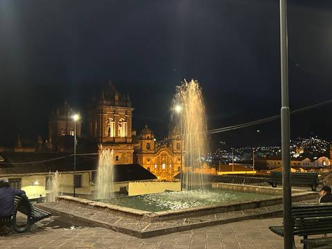       Night scene of Cusco cathedral and fountain jets glowing under warm lights.
  