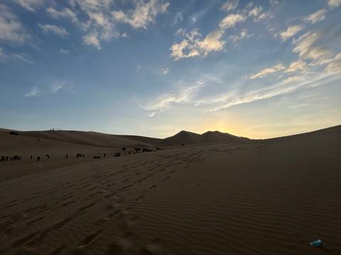       Sunset over desert dunes with footprints and a small group preparing for buggy rides.
  