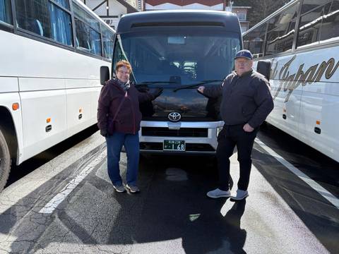       Two tourists pose beside a black Toyota minibus in a parking lot flanked by white tour buses.
  