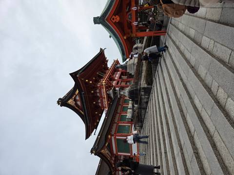       Vermilion-and-white shrine building atop stone steps with visitors ascending under cloudy skies.
  