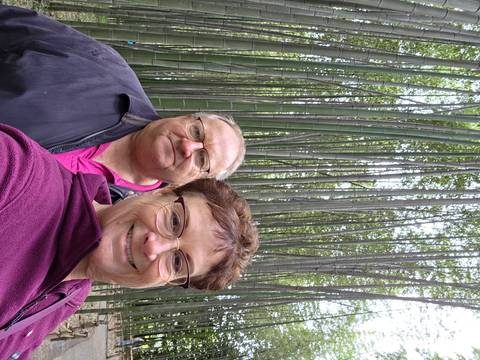       Selfie of two travelers in a dense bamboo grove with tall green stalks towering overhead.
  