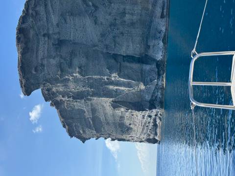       Dramatic volcanic sea cliffs of Santorini rise above clear blue Aegean waters viewed from a boat bow.
  