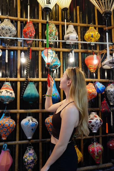       Young woman examines colourful lanterns in a shop, backlit by hanging lights.
  