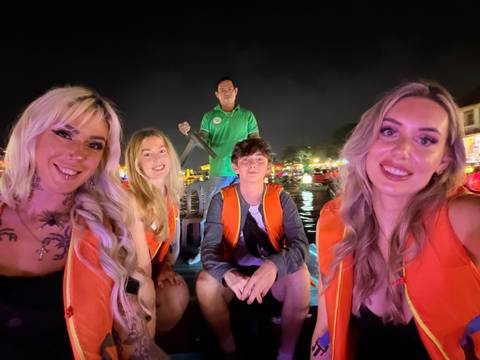       Group of friends in life jackets smiling on a small boat at night amid colourful river lanterns.
  