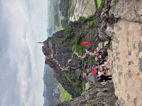       Steep stone stairway leading up a jagged limestone peak with red flags and many hikers visible, overlooking verdant rice fields.
  