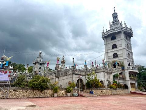       Grey stone castle-like shrine complex adorned with angel statues beneath dramatic storm clouds.
  