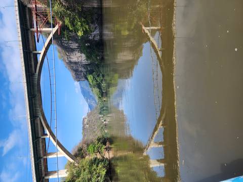       Tall bridge spanning a calm river with a reflection of the arch, framed by steep green cliffs and distant mountains.
  