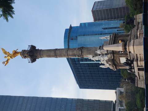       Monument of an angel-topped column surrounded by modern glass buildings under a pale blue sky.
  