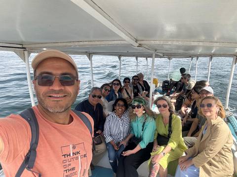       Selfie of a cheerful tour group seated on an open boat cruising a wide river
  