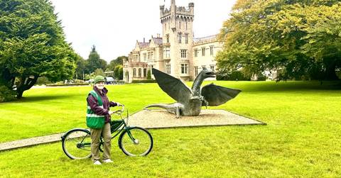       Cyclist posing beside a dragon sculpture on manicured castle grounds in Ireland.
  