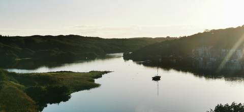       Serene estuary at dusk with a lone sailboat reflected in still water.
  