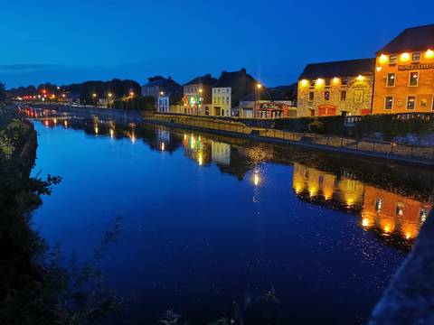       Riverside town in Ireland glowing with colourful lights and reflections in deep-blue twilight.
  