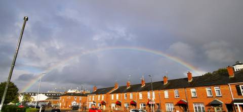       Full rainbow arching above rows of brick terraced houses under stormy clouds.
  