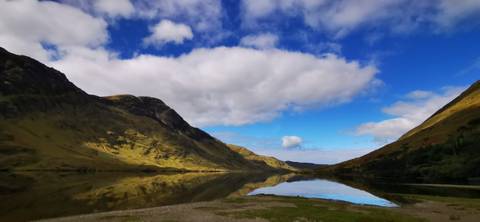       Mirror-like lake reflecting green mountains and a vivid blue cloud-speckled sky.
  