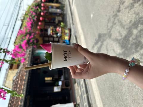       Hand holding a Mót Hoi An drink with bougainvillea and lantern-adorned street in background.
  