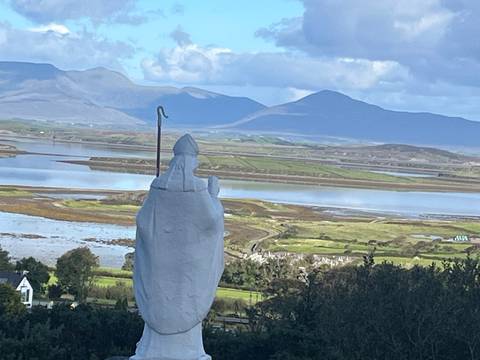       White statue of Saint Patrick overlooks an expansive tidal bay backed by mountains.
  