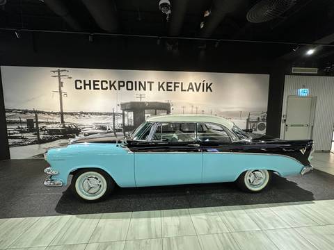       Vintage turquoise and black car displayed indoors with a large black-and-white backdrop reading “CHECKPOINT KEFLAVÍK”
  