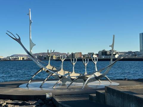       Sun Voyager steel sculpture by the waterfront with city skyline and distant mountains under a clear blue sky
  