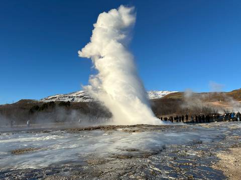       Spectacular geyser eruption shooting a white column of steam into a clear blue sky while tourists look on
  