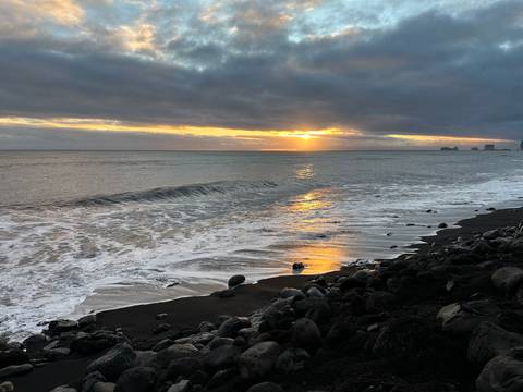       Golden sun setting over a black-sand beach with gentle waves and scattered rocks
  
