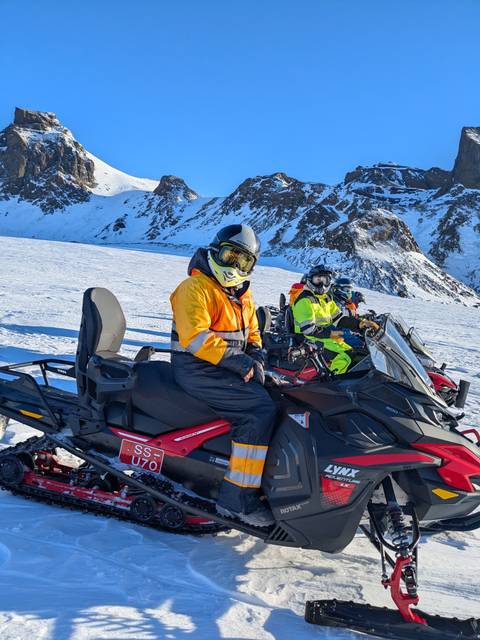       Riders in bright suits seated on snowmobiles atop a vast snowy glacier with craggy peaks in the background
  