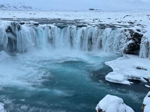       Wide turquoise pool at the base of a curtain-like icy waterfall surrounded by snow and ice
  