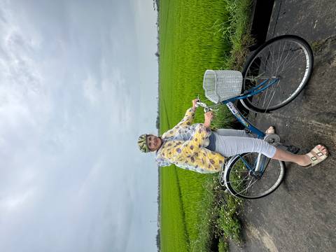       Woman in a rain poncho and helmet standing with her bicycle beside vivid green rice paddies under a grey sky
  