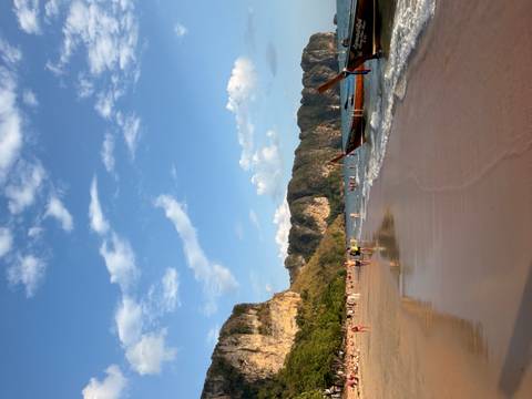       Sandy beach at sunset with long-tail boat ashore and people wading in gentle waves under blue sky.
  