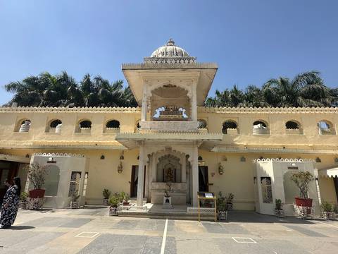       Elegant yellow-stone temple courtyard with carved arches, domed pavilion and palm trees under a clear sky.
  