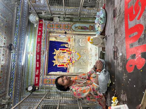       Young priest or devotee sits cross-legged inside a richly decorated Hindu shrine before a deity statue.
  