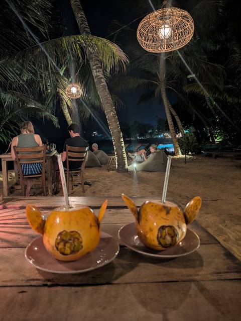       Nighttime beach restaurant with guests dining on sand under palm trees; coconut cocktails in foreground.
  