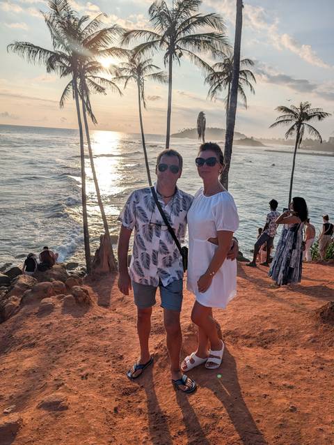       Couple poses on a red-earth headland lined with leaning palm trees overlooking the ocean at golden sunset.
  