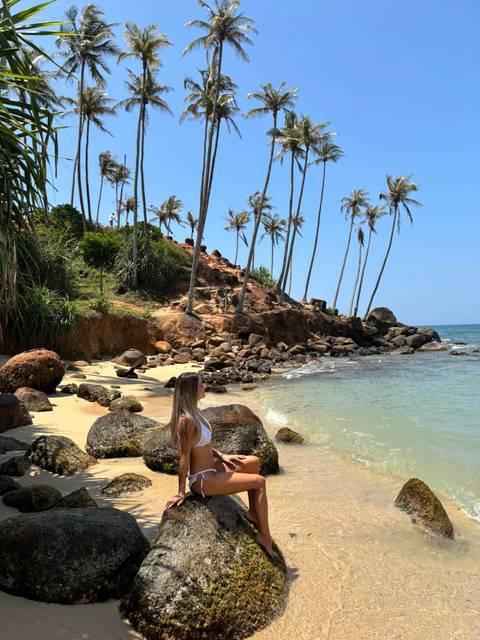       Woman relaxing on a secluded sandy cove with rock formations and leaning palm trees
  
