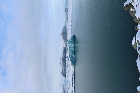       Serene glacial lagoon with a blue iceberg floating before snow-covered mountains under a pale sky.
  