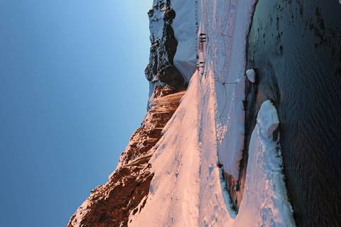      Sunlit waterfall cascading over a snow-blanketed cliff glowing pink in low winter light, with people visible at the base.
  