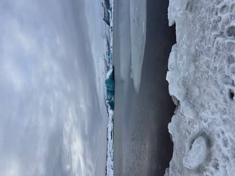       Calm winter lagoon with floating icebergs beneath overcast grey skies and a snowy shore.
  