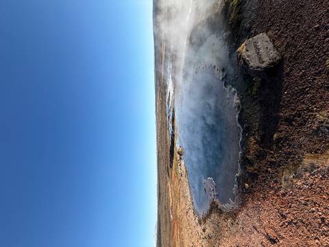       Steaming geothermal pool amid a barren plain under a clear blue sky.
  