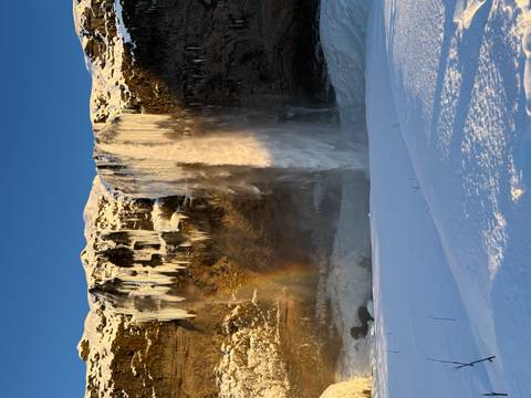       Tall Seljalandsfoss waterfall framed by icicles and a faint rainbow, dropping into a snowy meadow.
  
