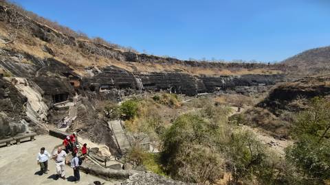       Panoramic view across an arid valley revealing a row of rock-cut Ajanta caves in a cliffside, with tourists on a walkway.
  