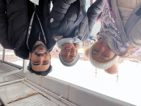       Three friends bundled in winter gear smile on a Bosphorus ferry with Istanbul mosques behind.
  