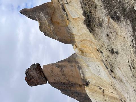       Balanced mushroom-shaped rock looming above pale striped sandstone slopes under a grey sky.
  