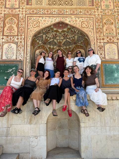       Large group of women posing happily on decorative steps of an Indian palace interior.
  