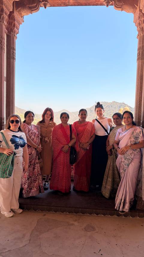       Tourists and local women standing together with a hilly fort backdrop in Rajasthan.
  