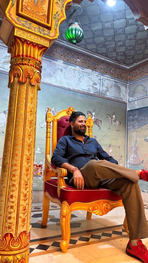       Man seated on an ornate golden throne chair against a backdrop of traditional Indian miniature paintings.
  