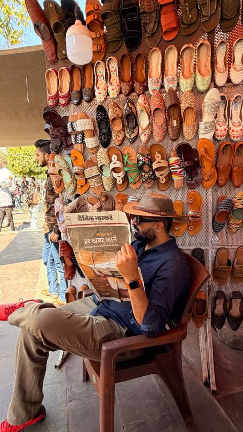       Man reading a Hindi newspaper at a vibrant street stall selling colourful sandals in India.
  