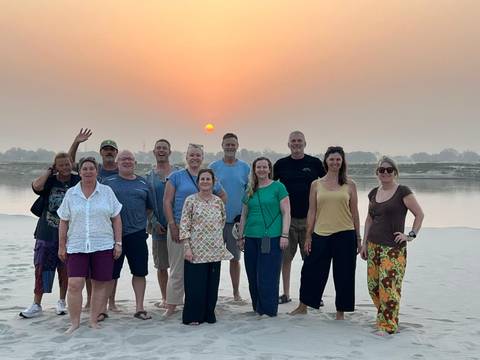       Group of travellers posing at sunset on a sandy riverbank with the sun over the Ganges.
  