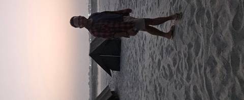       Man walking near tents on a dimly lit sandy riverside camp at dusk.
  